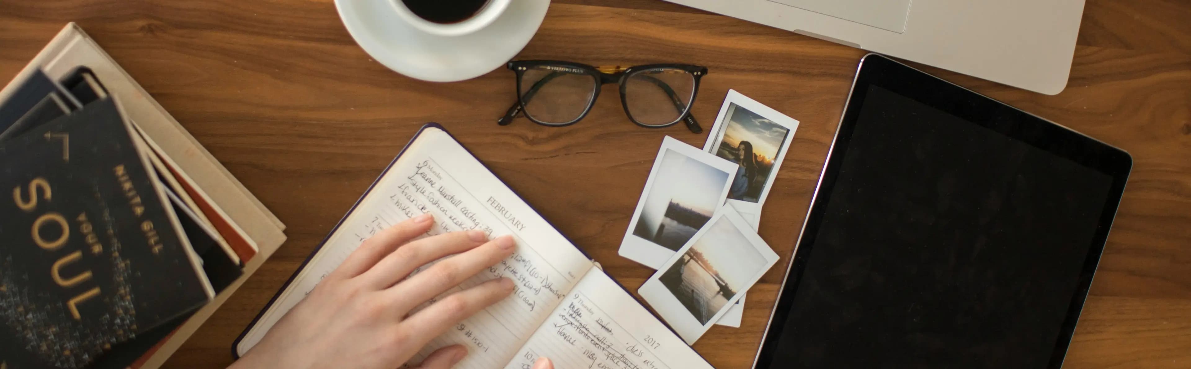A desk seen from above: an open notebook being written in, a cup of black coffee, reading glasses, instant photographs, a tablet and a laptop.